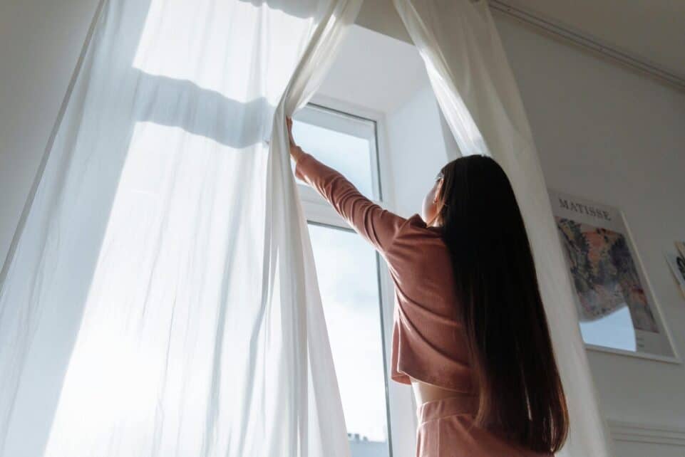 Woman opens white curtains in bright room, revealing a sunny day outside.