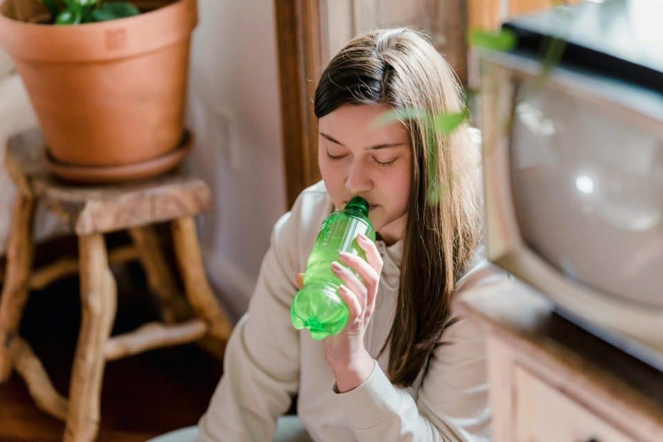 Tranquil woman with brown hair in casual clothes drinking soda from green bottle at home in daylight