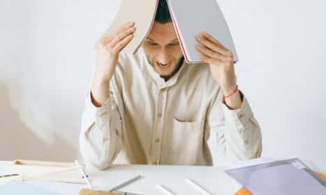 A stressed adult male worker sits at a desk with open notebooks, exhibiting signs of frustration and burnout.