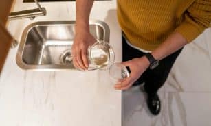 A person wearing a yellow sweater pours water into a glass at a kitchen sink.