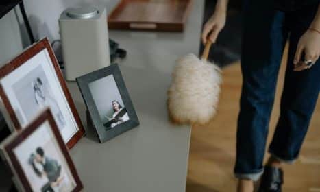 A woman using a feather duster to clean picture frames on shelves indoors.