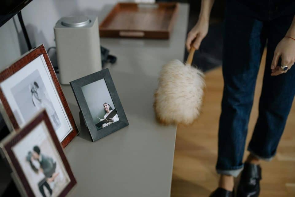 A woman using a feather duster to clean picture frames on shelves indoors.