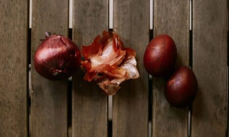 Close-up of a red onion, peels, and red eggs on a wooden table surface.