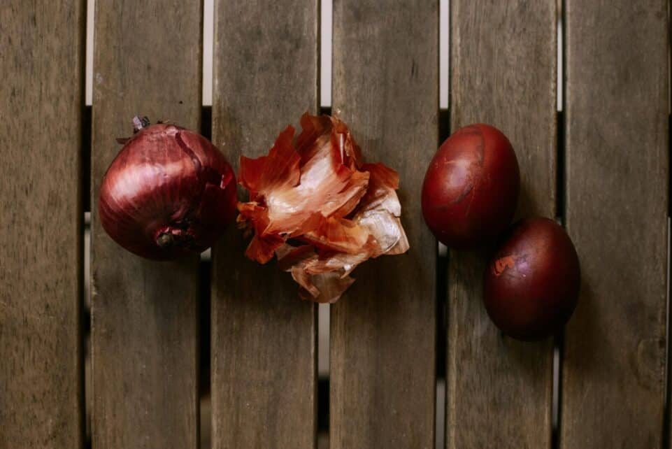 Close-up of a red onion, peels, and red eggs on a wooden table surface.