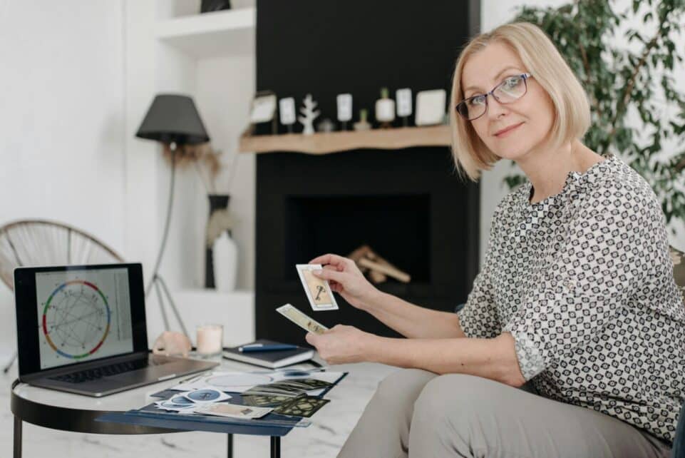 Woman with tarot cards using laptop for astrology reading in cozy living room.