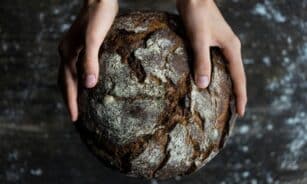 Close-up of hands holding a rustic loaf of whole wheat bread with a dark, textured crust.