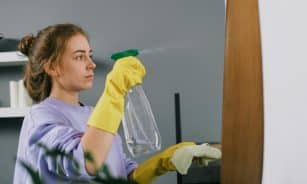 Side view of young calm female in protective latex gloves sprinkling liquid detergent from plastic bottle on mirror in light room