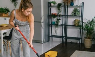 A young woman in a jumpsuit mopping a modern kitchen floor with a red mop, surrounded by plants.