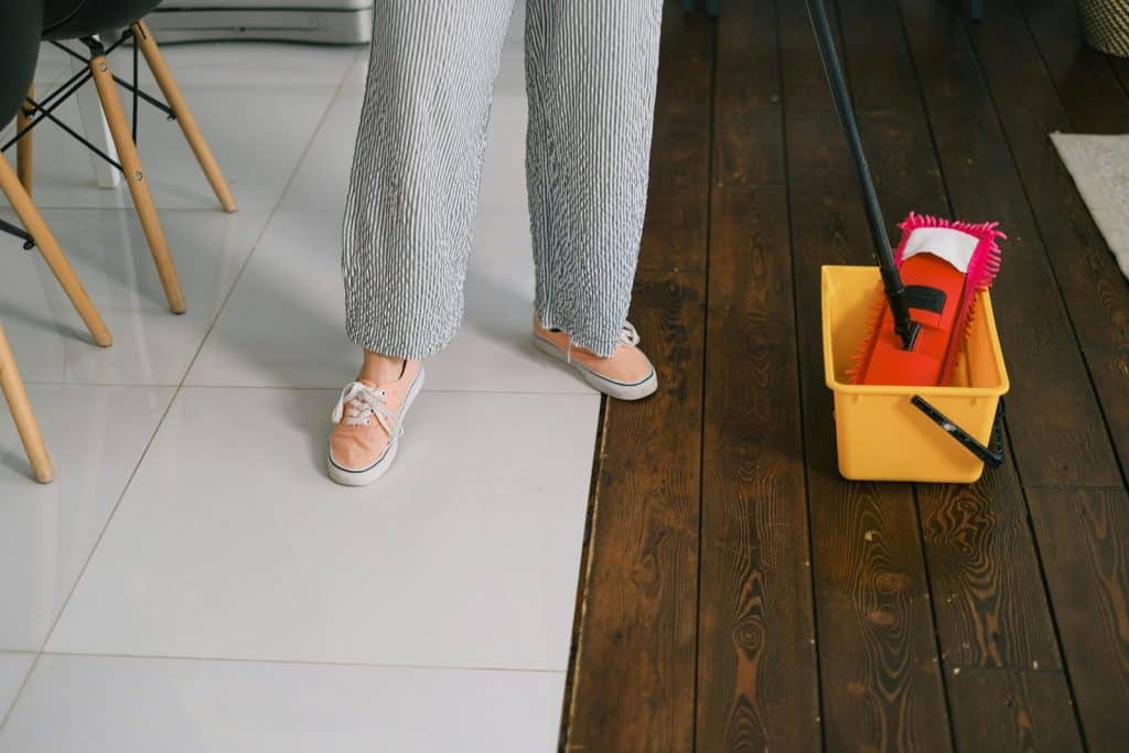 Feet of a person cleaning indoors with mop and bucket on wooden floor.