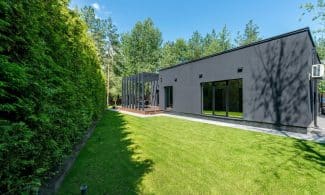 Sleek black modern house with large windows and vibrant green lawn surrounded by dense trees.