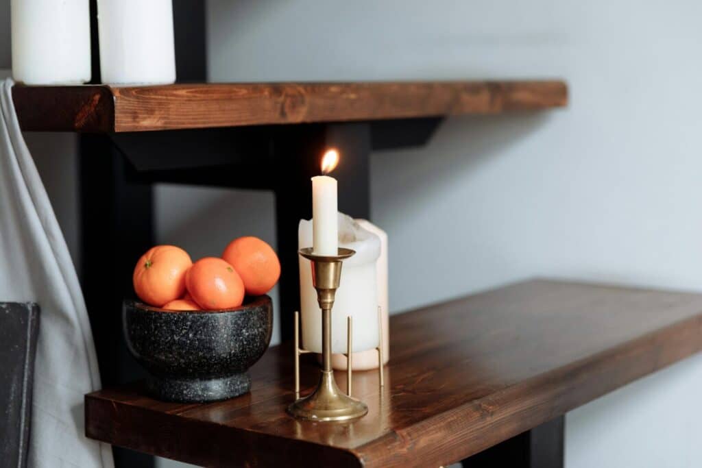 Warm, inviting scene with a lit candle and bowl of oranges on a wooden shelf.
