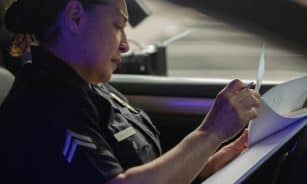 A female police officer sits in a patrol car, focused on writing a report during daylight.