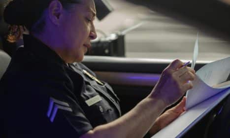 A female police officer sits in a patrol car, focused on writing a report during daylight.