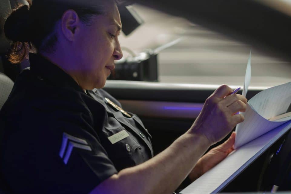 A female police officer sits in a patrol car, focused on writing a report during daylight.