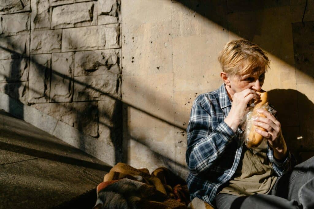 A senior man sitting against a wall, eating bread, highlighting homelessness.