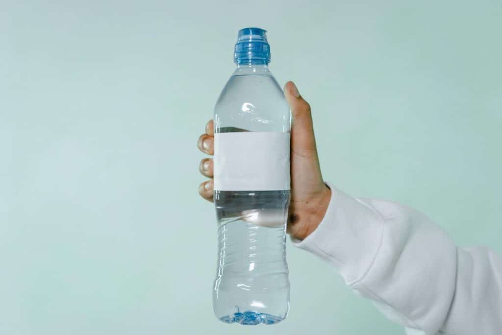 A hand holding a clear plastic water bottle with blank label on a soft green background.