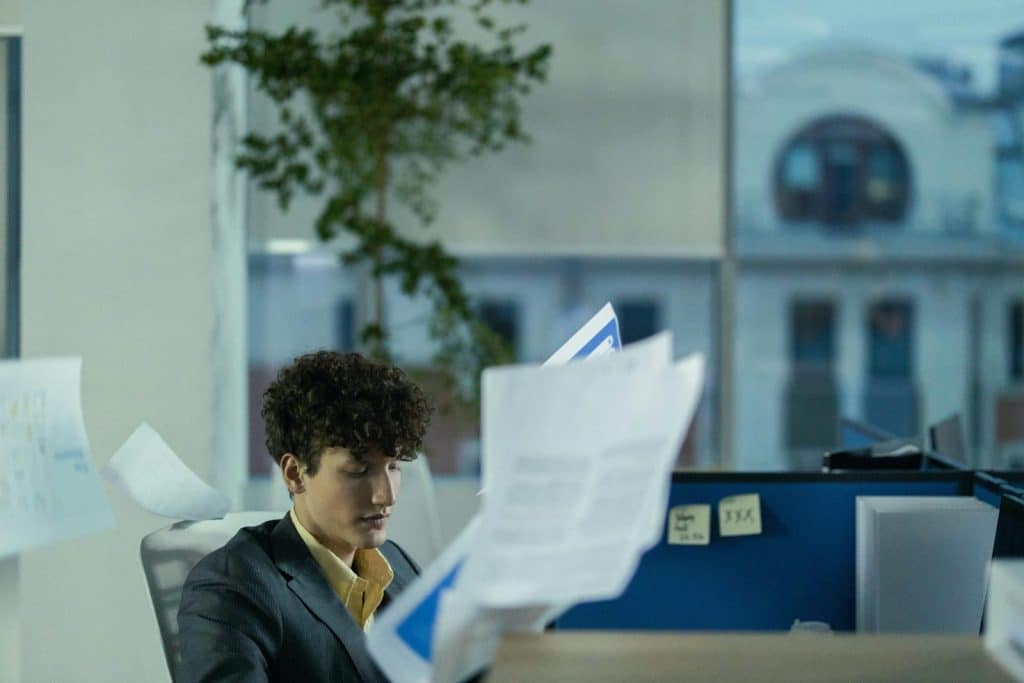 Young man in office cubicle tossing papers, illustrating busy work environment.
