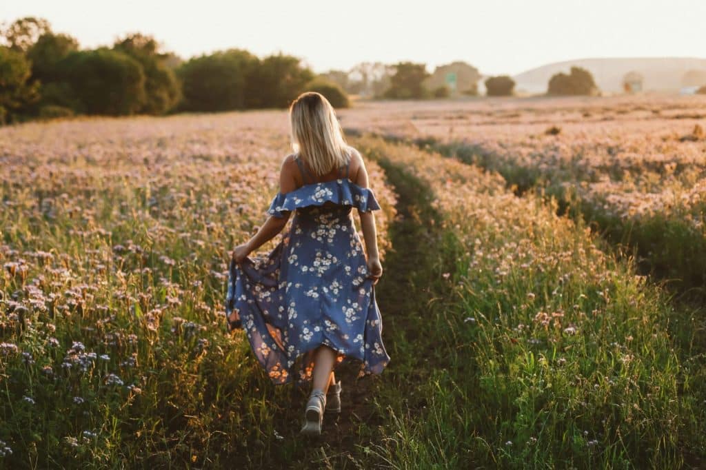 A woman walking in a floral dress through a bright flower field during sunset, showcasing tranquility and nature.