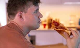 A man savors a slice of pepperoni pizza in a warmly lit indoor setting, side view emphasized.