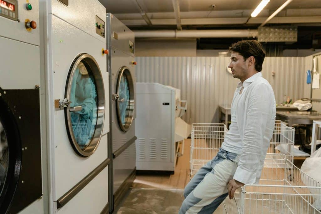A man sits in a laundromat next to industrial washing machines, waiting for his laundry.