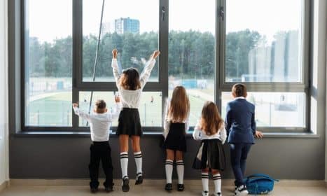A group of school children in uniform stand by a large window, looking outside.