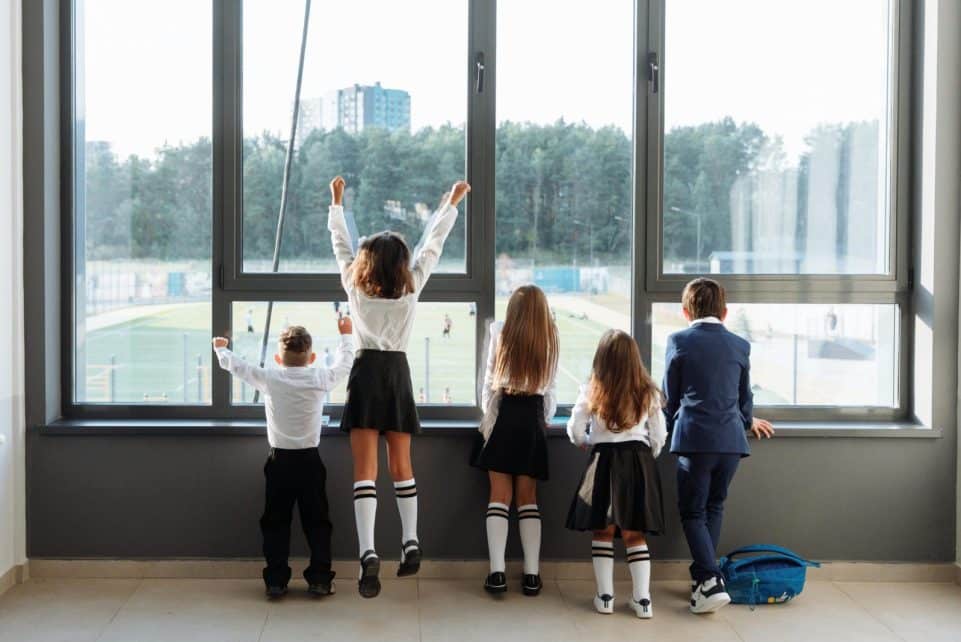 A group of school children in uniform stand by a large window, looking outside.