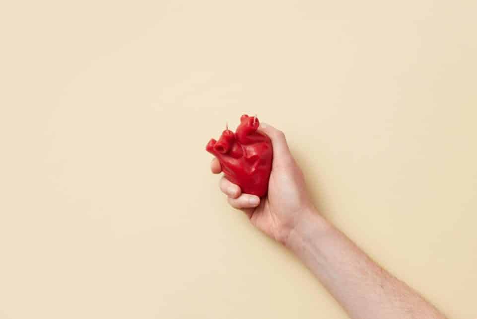 A realistic red anatomical heart model held firmly in a hand against a beige background.