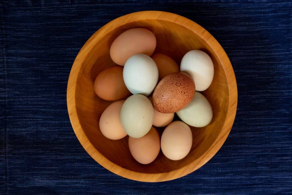 A variety of colorful chicken eggs arranged in a wooden bowl on a dark blue background.