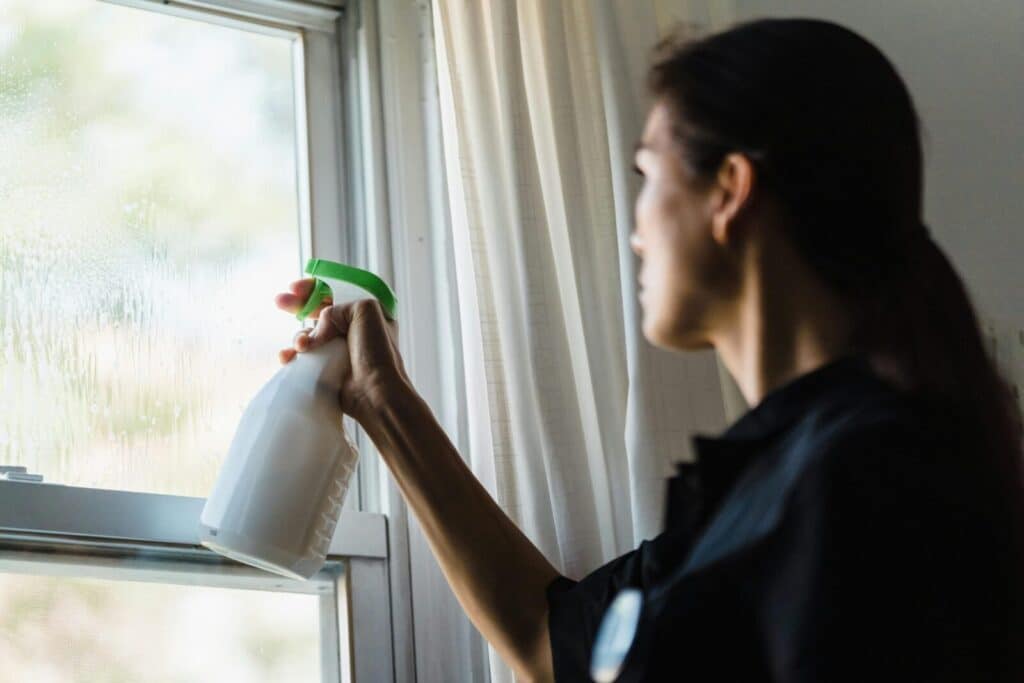 A woman using a spray bottle to clean a glass window inside a room, captured in a close-up shot.
