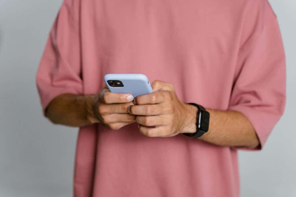 Adult male in pink t-shirt using smartphone; focus on hands and phone.
