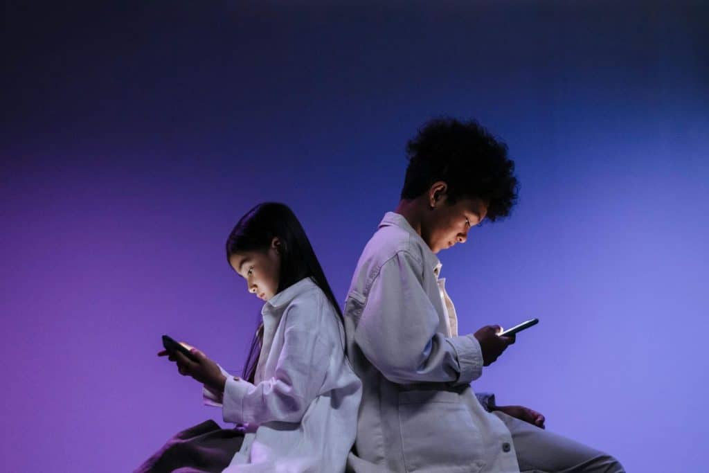 Two teenagers sitting back to back, focused on smartphones with vivid lighting.