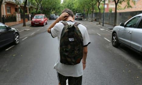 boy wearing white shirt and black shorts carrying backpack standing on black concrete road between vehicles and trees during daytime