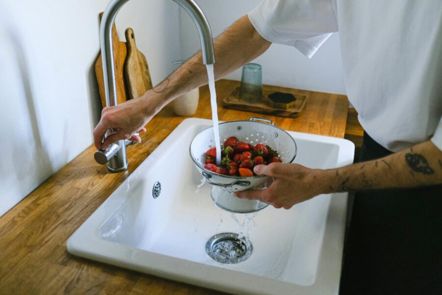 A person washes fresh strawberries in a colander under a kitchen faucet.