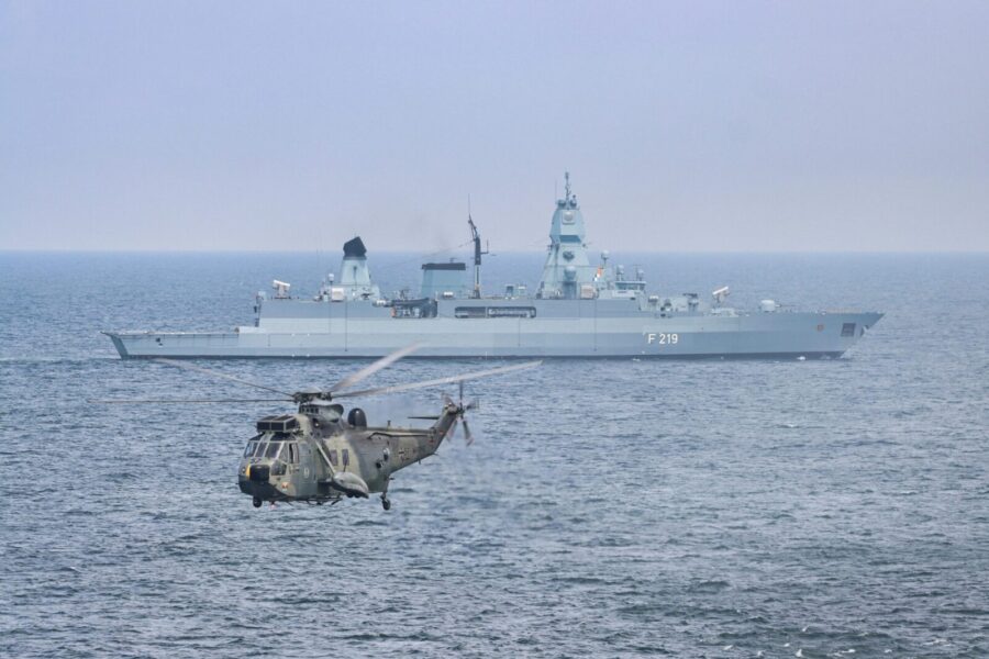 A military helicopter flies over the sea near a German warship, F219.