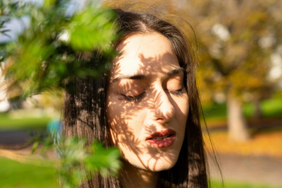 A woman with eyes closed in sunlight, leaf shadows cast on her face, creating a serene and artistic portrait.