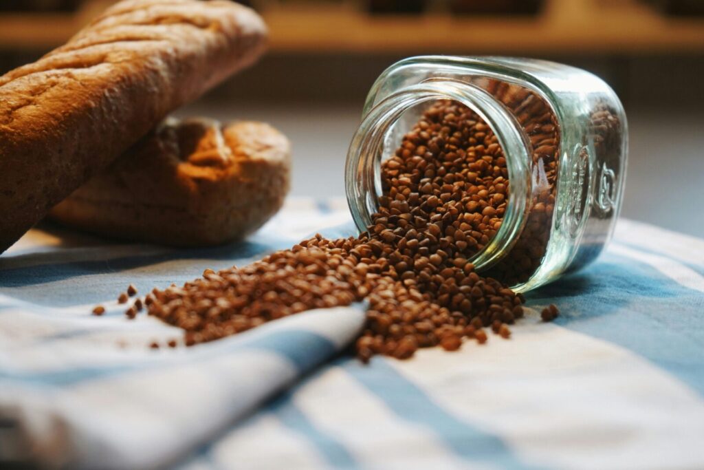 A rustic scene with buckwheat spilling from a jar next to fresh bread on a checkered cloth.