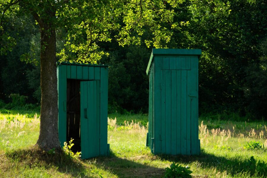 Two green wooden huts in a sunny rural setting surrounded by lush greenery.