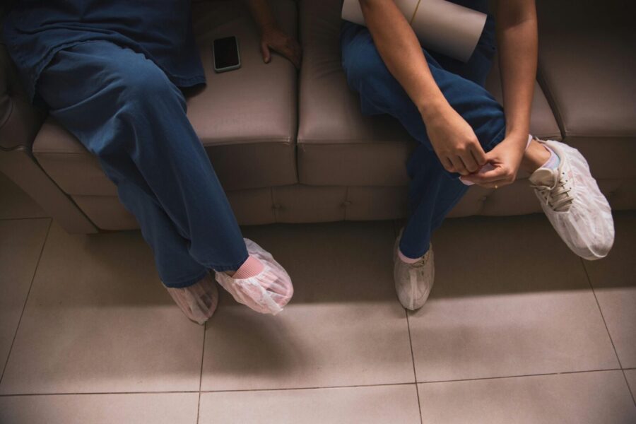 Two healthcare workers sitting on a sofa wearing scrubs and protective shoe covers.