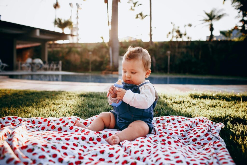 A cute baby sitting on a blanket in a sunny backyard, holding a bottle and enjoying a picnic.