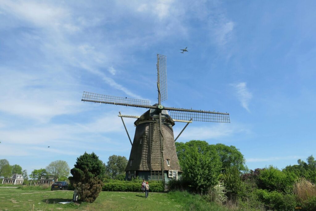 A classic Dutch windmill stands tall in a lush green field under a clear blue sky with an airplane flying above.