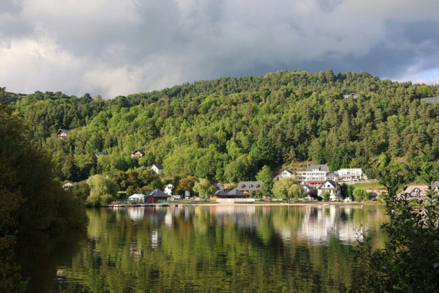 Charming village houses reflected in a serene forest-surrounded lake under a cloudy sky.