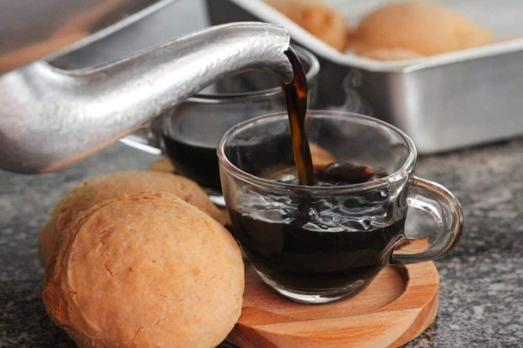 Steaming coffee poured into glass cup beside fresh bread rolls on wooden platter.
