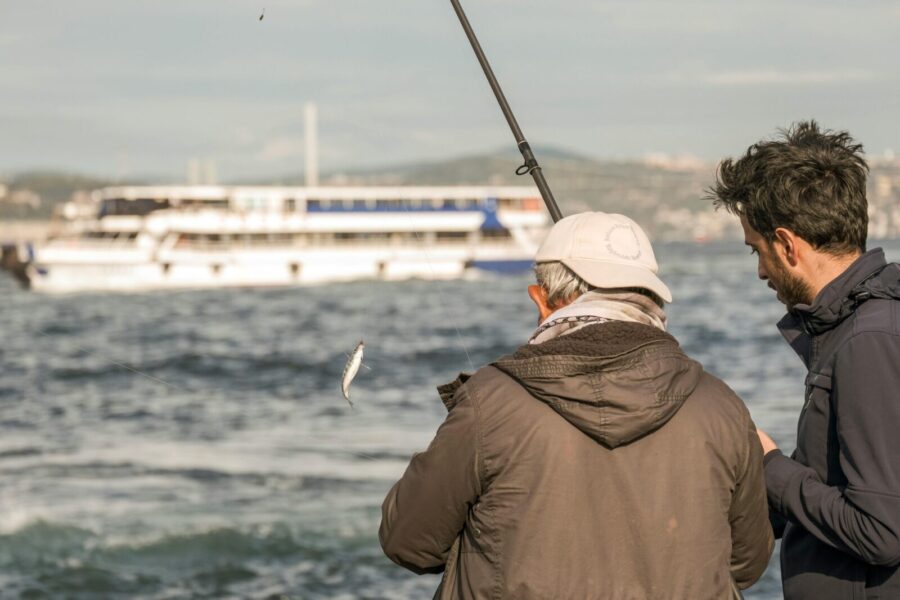 Two men fish near the sea as a ferry boat passes in the background, creating a serene waterfront scene.
