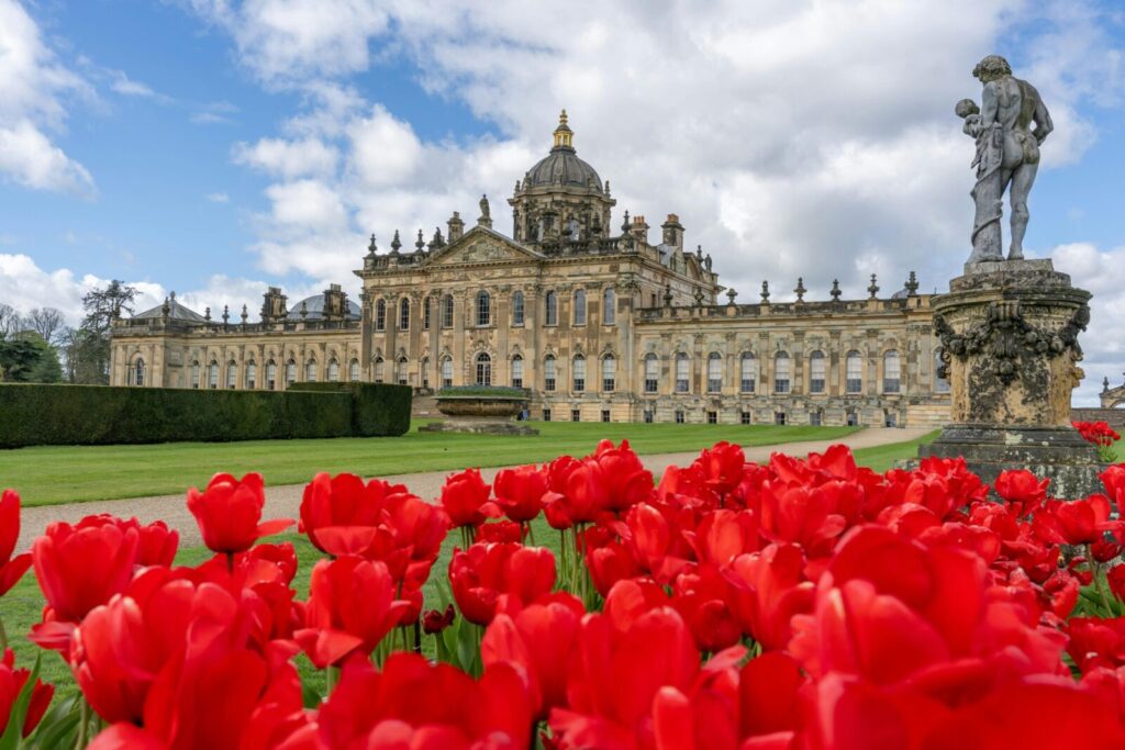Vibrant red tulips foreground Castle Howard's grand architecture under a partly cloudy sky.