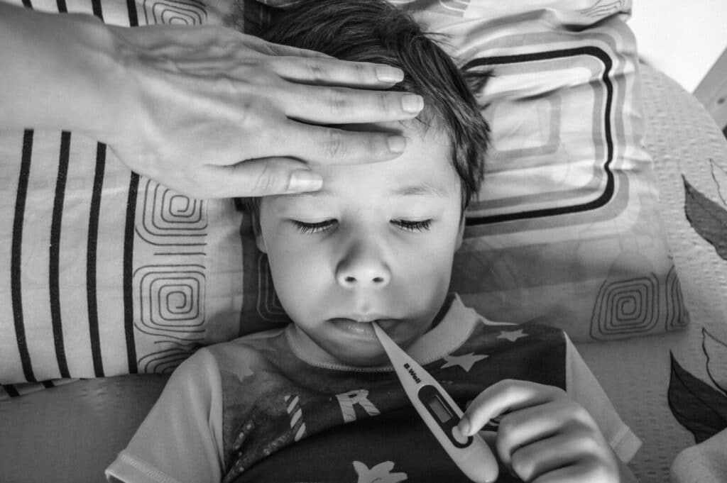 Black and white from above of unrecognizable person touching forehead of unemotional child in casual clothing holding electronic thermometer in mouth