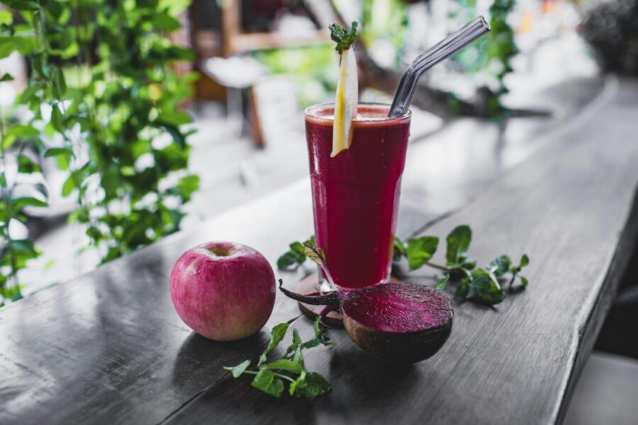 Glass of fresh cold healthy smoothie placed on wooden table on terrace of cafe with whole apple and halved beetroot
