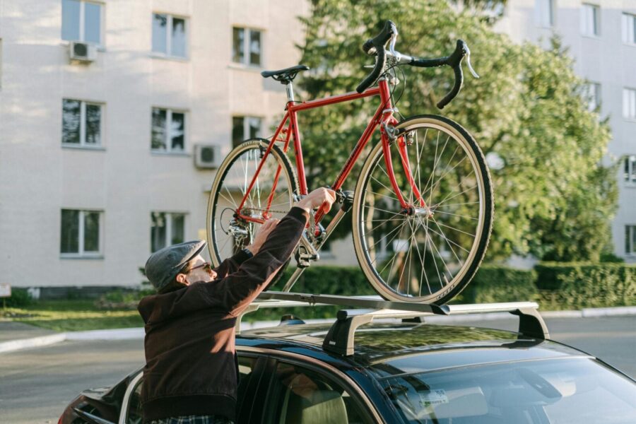 A man secures a red bicycle on top of a car's roof rack in an urban setting.