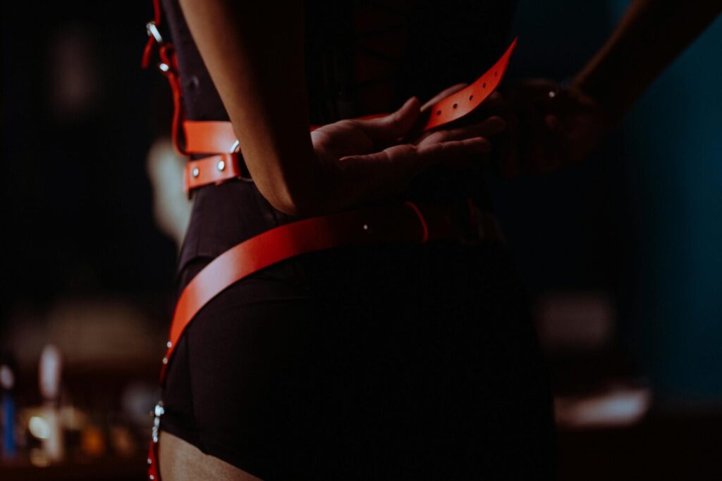 Back view of crop unrecognizable female in black corset and panties fastening leather harness in dressing room