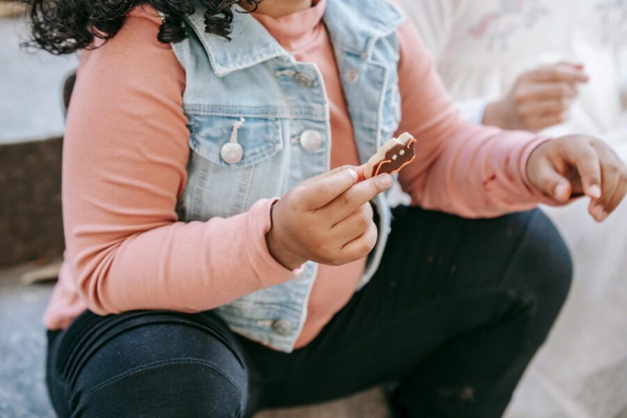 Crop anonymous ethnic girl in casual wear sitting on staircase and eating bat shaped gingerbread at Halloween celebration