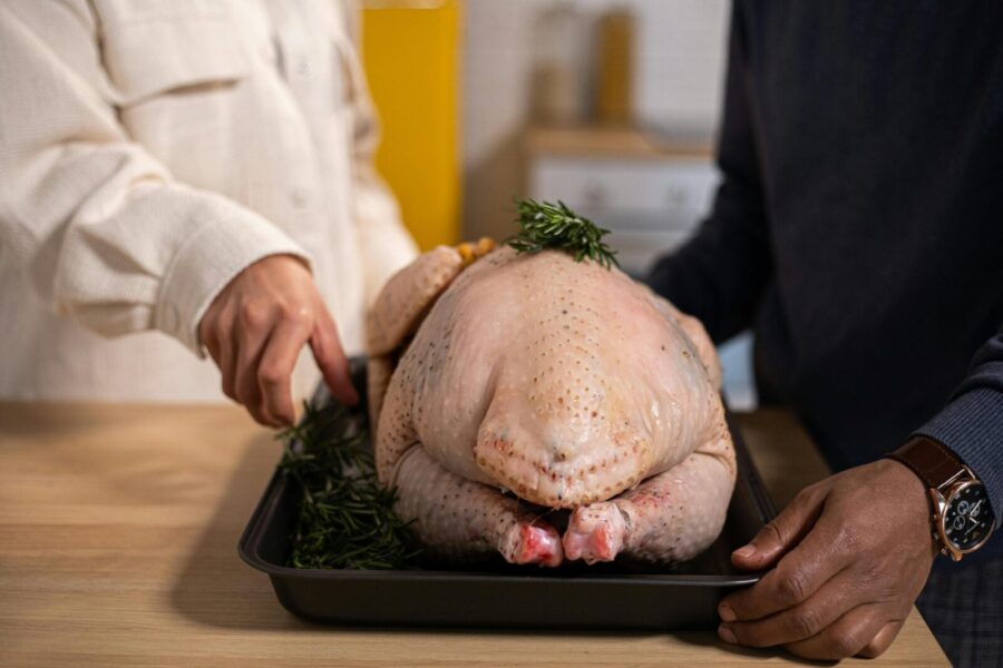 Unrecognizable African American couple standing at wooden counter with tray of uncooked whole turkey topped with rosemary in kitchen on blurred background